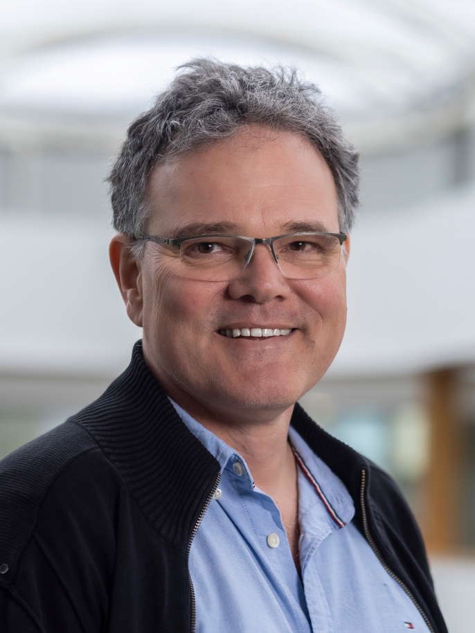 This picture shows a portrait of a male physicist with glasses, dark hair, wearing smart casual clothes.